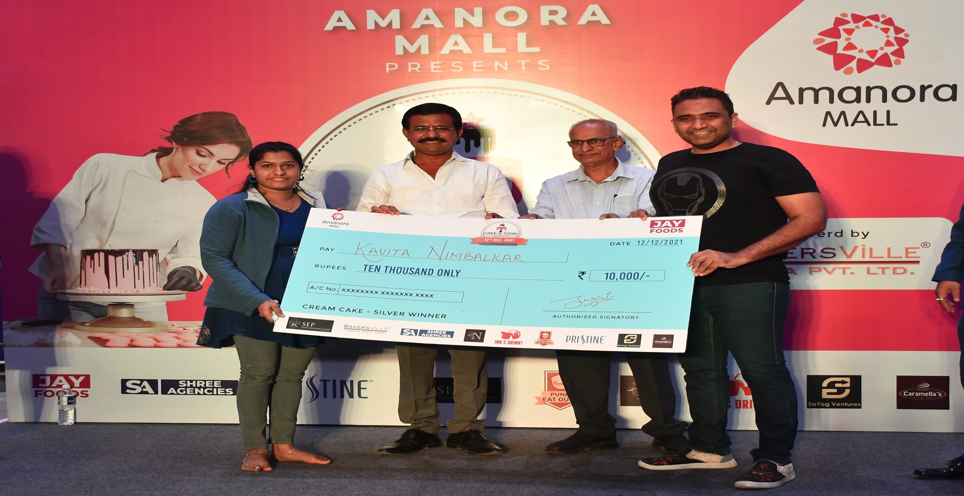 Cake-A-Thon Season 3 prize presentation: Kavita Nimbalkar and others on stage holding an oversized cheque for Rs 10,000, marked "Cream Cake - Silver Winner", in front of an Amanora Mall backdrop.