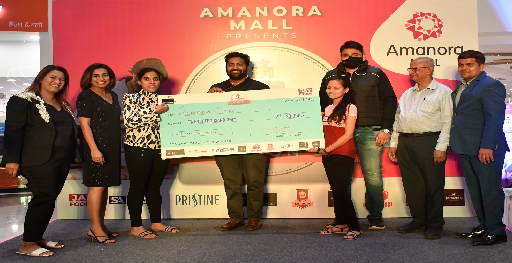 Cake-A-Thon Season 3 prize presentation: Aishwarya Gire and others on stage holding an oversized cheque for Rs 20,000, marked "Fondant Cake - Gold Winner", in front of an Amanora Mall backdrop.