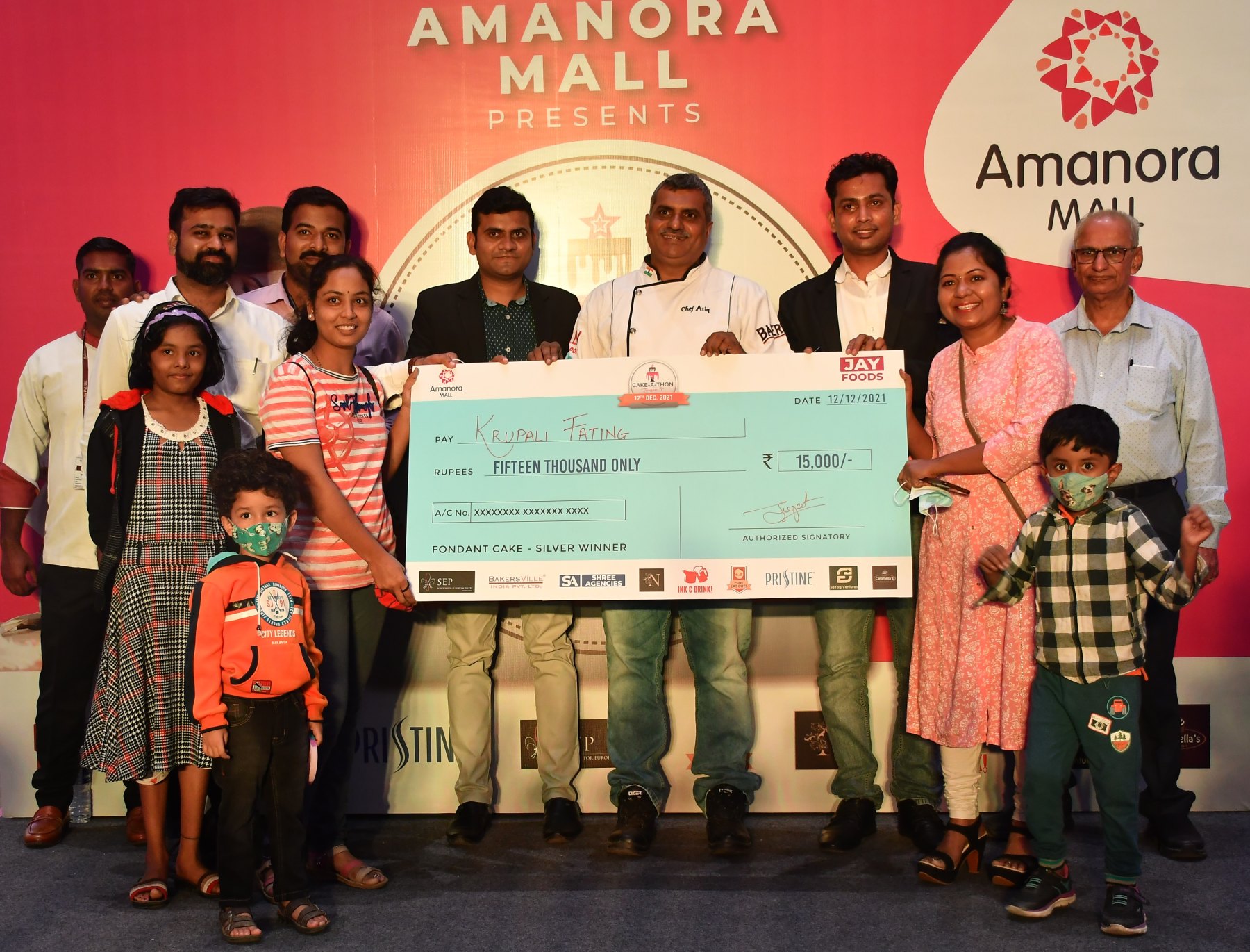 Cake-A-Thon Season 3 prize presentation: Krupali Fating and others on stage holding an oversized cheque for Rs 15,000, marked "Fondant Cake - Silver Winner", in front of an Amanora Mall backdrop.