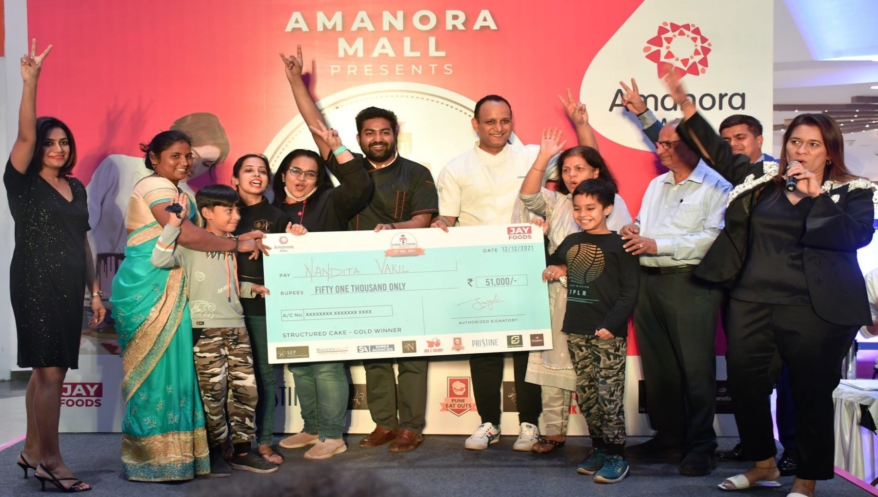 Cake-A-Thon Season 3 prize presentation: Nandita Vakil and others on stage holding an oversized cheque for Rs 51,000, marked "Structured Cake - Gold Winner", in front of an Amanora Mall backdrop.