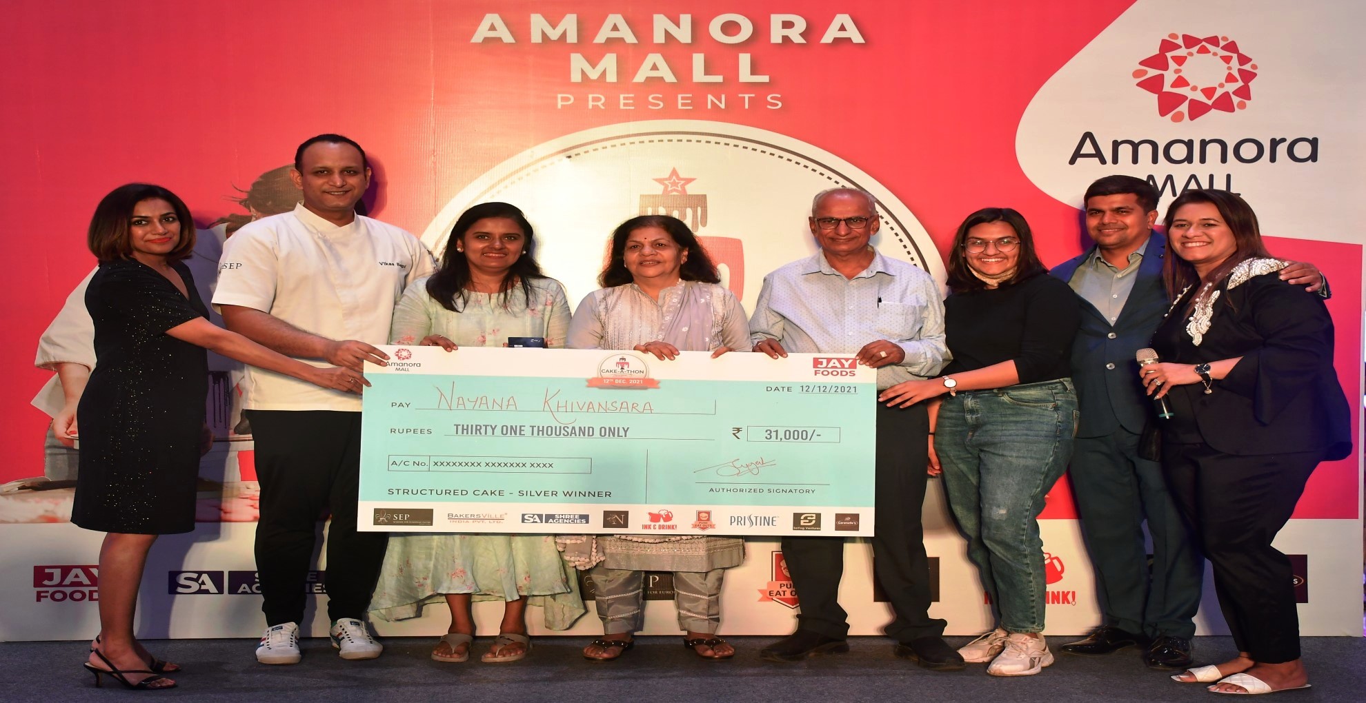 Cake-A-Thon Season 3 prize presentation: Nayana Khivansara and others on stage holding an oversized cheque for Rs 31,000, marked "Structured Cake - Silver Winner", in front of an Amanora Mall backdrop.